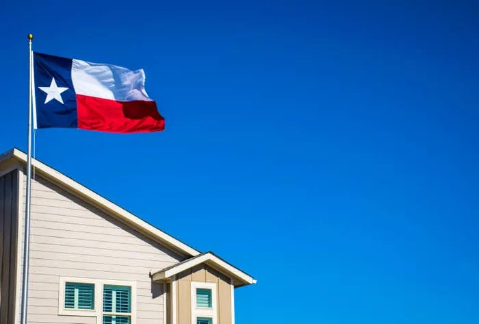 Una bandera del estado de Texas ondea en un mástil junto a una casa con un cielo azul claro en el fondo