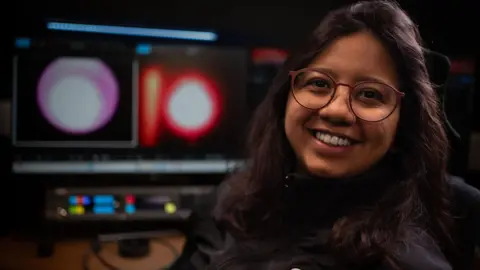 Tony Jolliffe/BBC News Una mujer, Verónica Viera, está sentada frente a dos grandes monitores de computadora en una habitación oscura. Las pantallas muestran dos imágenes de una forma circular blanca y roja: se trata de un plasma brillante en el horno espacial. Verónica tiene el pelo largo y castaño, usa gafas y una chaqueta negra.