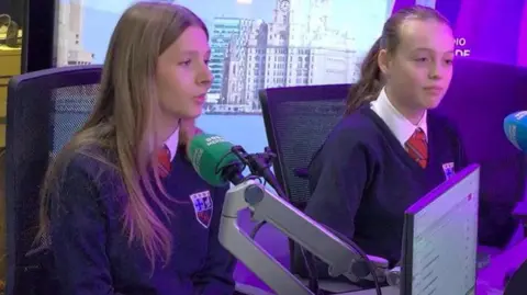 BBC Dos niñas con uniformes escolares están en un estudio de radio frente a micrófonos. 