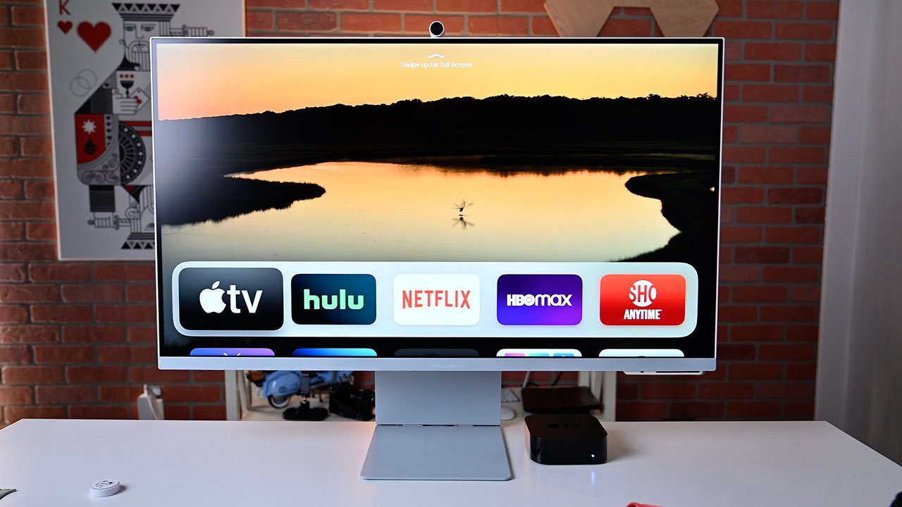 Una pantalla de computadora M8 que muestra íconos de servicios de transmisión contra una escena de la naturaleza, sobre un escritorio blanco, con una pared de ladrillos y una gran baraja de cartas al fondo.