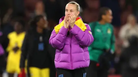 PA Media Una mujer con cabello rubio recogido hacia atrás y vestida con un abrigo acolchado morado y amarillo y pantalones cortos ingleses está llamando a la puerta. Ella está parada en un campo de fútbol.