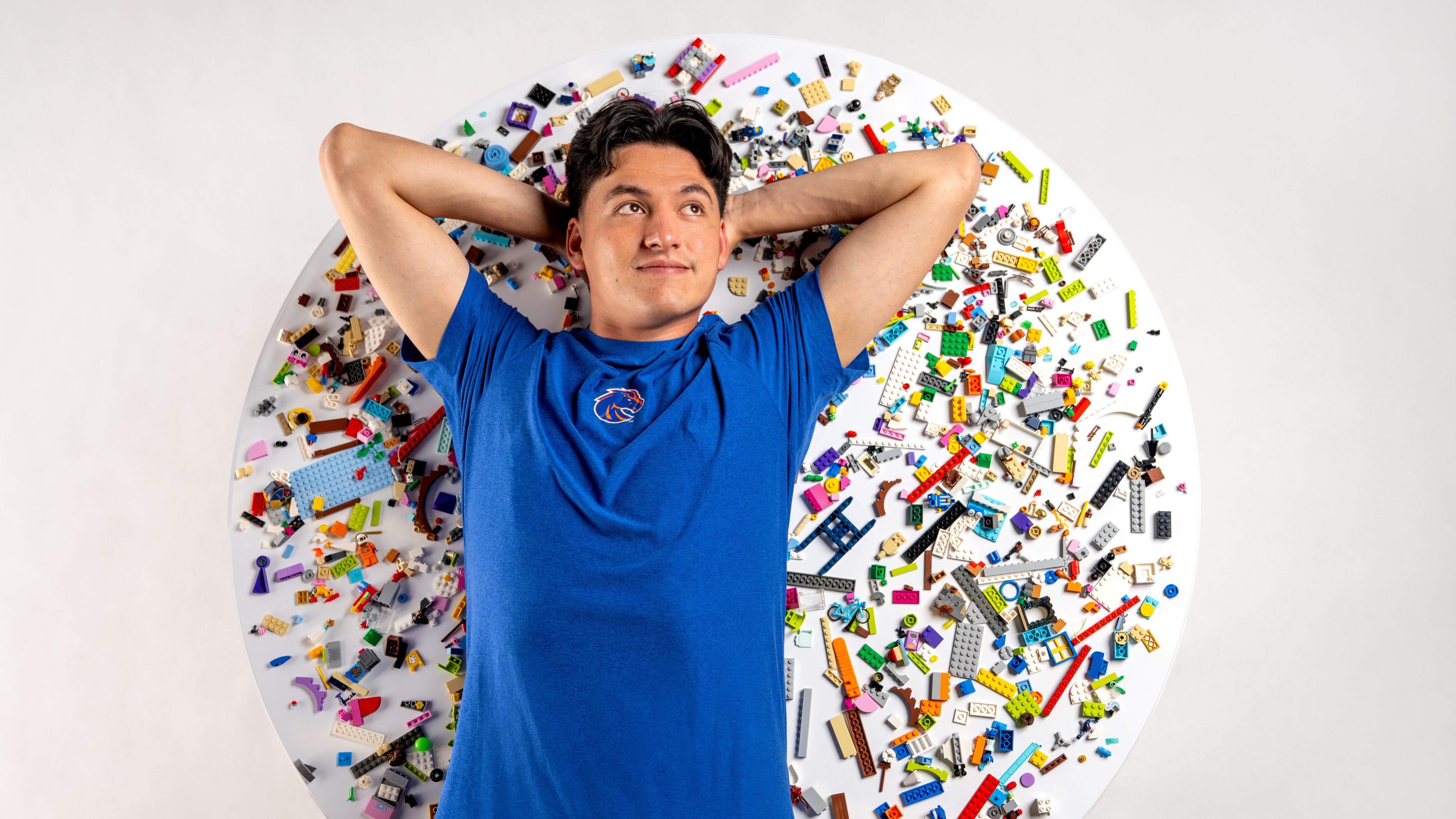 González, vestido con una camiseta azul de Boise State, está de pie sobre una mesa llena de ladrillos LEGO.