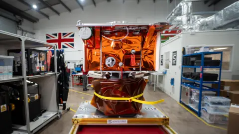 Tony Jolliffe/BBC News Una maqueta a tamaño real del satélite de la fábrica espacial se encuentra en una mesa de trabajo dentro de la sede de Space Forge en Cardiff. Tiene forma cuadrada, aproximadamente del tamaño de un microondas, y está cubierto con una lámina naranja reflectante con varios componentes más pequeños adjuntos. El taller al fondo tiene techos altos y estantes llenos de equipos y cajas de almacenamiento. Una gran bandera Union Jack cuelga de la pared.