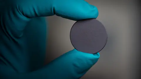 Tony Jolliffe/ BBC News Un primer plano de una mano enguantada que sostiene un pequeño disco gris circular, que es una esfera de material utilizado en semiconductores. El guante es azul y el disco se sostiene entre dos dedos. 