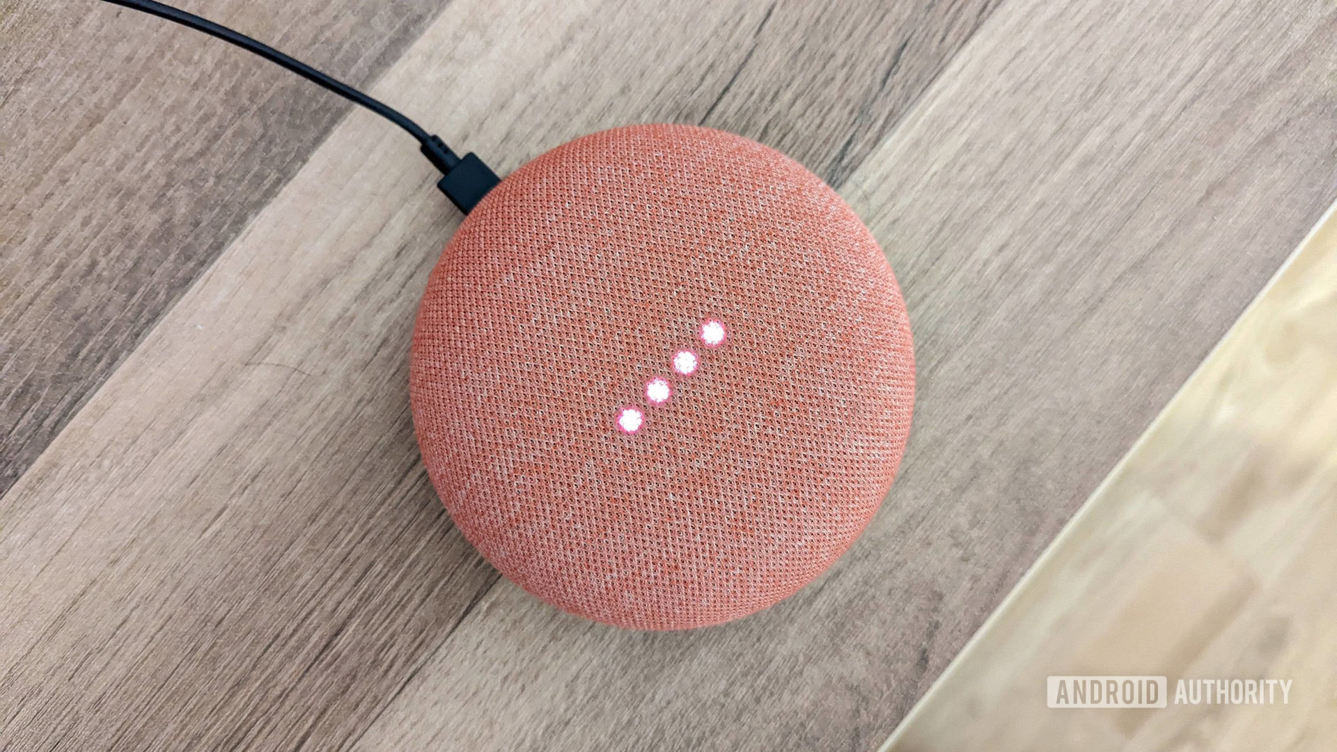Luces coralinas de Google Home Mini encendidas Google Home Mini en color coral con las luces encendidas, sobre fondo de madera