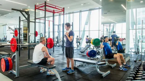 Getty Images La gente entrena en lo que parece un gimnasio comercial. Un hombre está sentado en un banco de pesas charlando con un hombre frente a él. En la esquina derecha, un hombre levanta pesas con ambos brazos. Al fondo se puede ver a mujeres usando motores de bicicletas. 