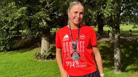 Leah Walsh Leah sonríe mirando a la cámara. Ella se para frente a los árboles y la vegetación y viste una camiseta roja de finalista de la media maratón de Cardiff, una medalla negra y pantalones cortos negros para correr. Leah tiene el pelo rubio recogido en una coleta alta. 