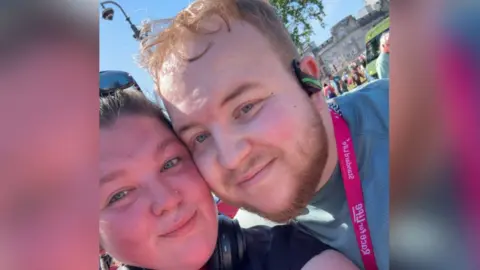 Richard Gallimore Un selfie en primer plano de Richard con su novia, Sophie, en el evento de carreras Race for Life. Sophie, a la izquierda, tiene cabello castaño y usa gafas de sol en la parte superior de la cabeza. Richard está de pie junto a Sophie con su mejilla tocando la de ella. Viste una camisa verde, una medalla rosa, auriculares y tiene el pelo rubio corto. 