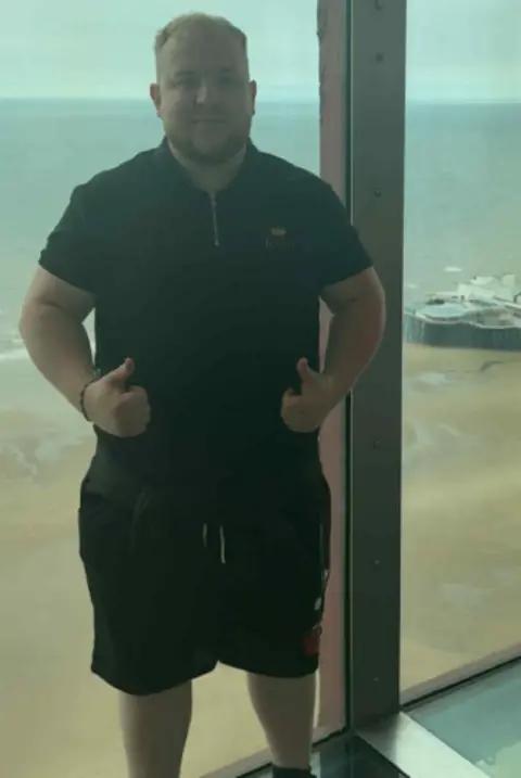 Richard Gallimore Richard parado frente a una ventana que muestra una vista de la playa. Está mirando a la cámara con el pulgar hacia arriba y lleva una camiseta polo negra con pantalones cortos negros. 