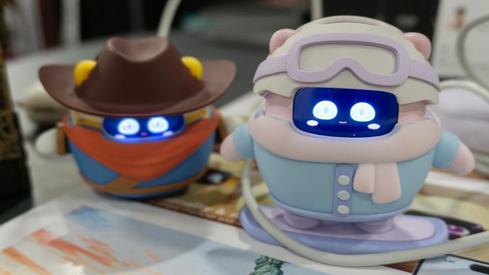 Se ven dos dispositivos Sweekar sobre una mesa, uno con un traje de snowboard rosa y azul y el otro (detrás) con un sombrero y un traje de vaquero.