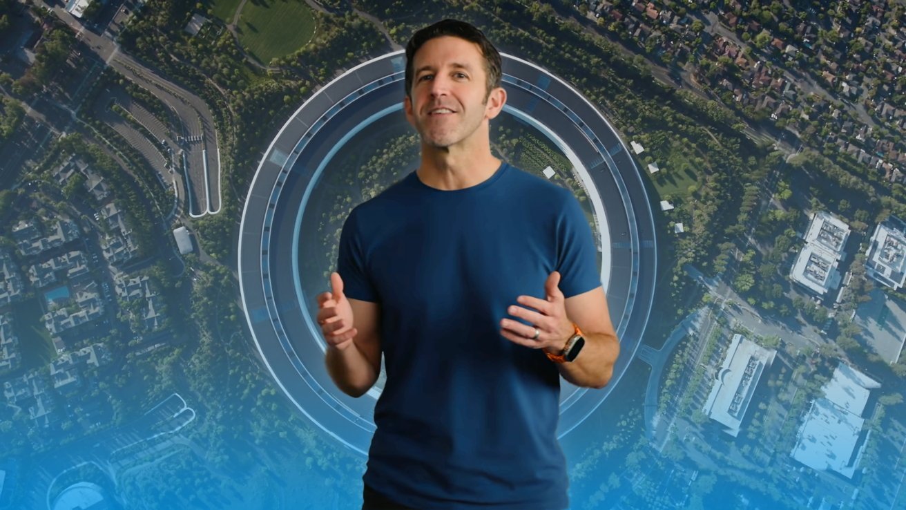 Un hombre con una camisa azul habla con gestos con las manos, parado frente a una vista aérea de un gran campus de oficinas circular rodeado de vegetación y edificios de la ciudad cercanos.