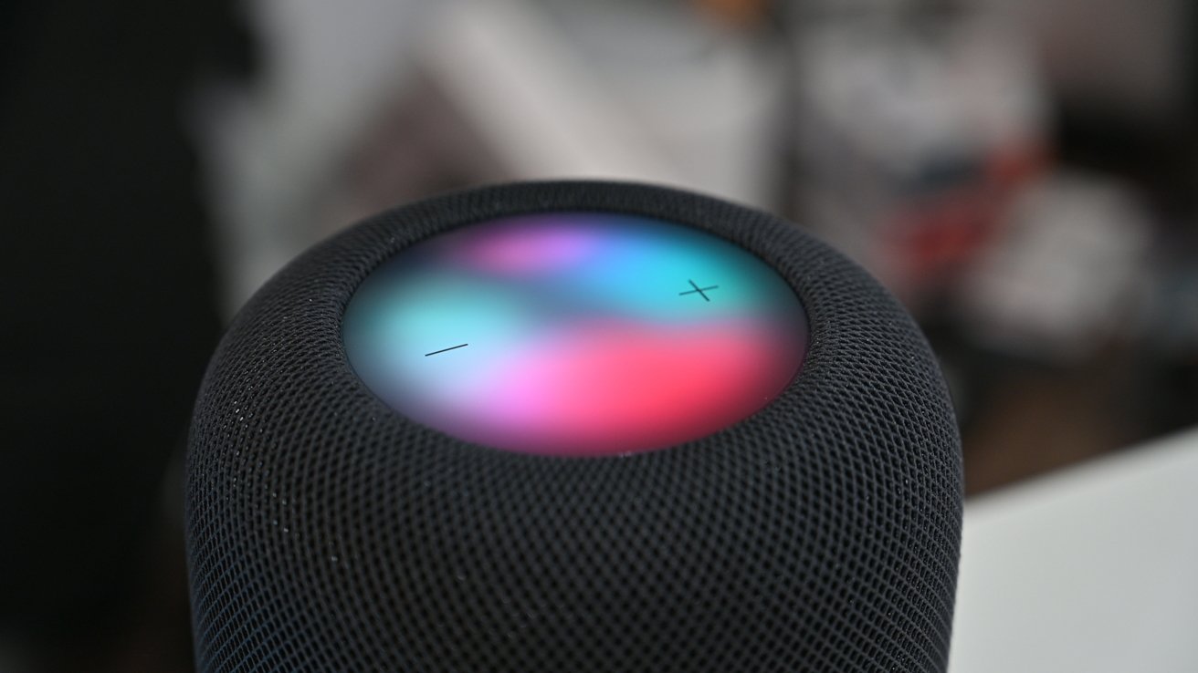 Primer plano de un HomePod negro con lados de malla y una superficie táctil multicolor brillante en la parte superior, que muestra símbolos de volumen más y menos