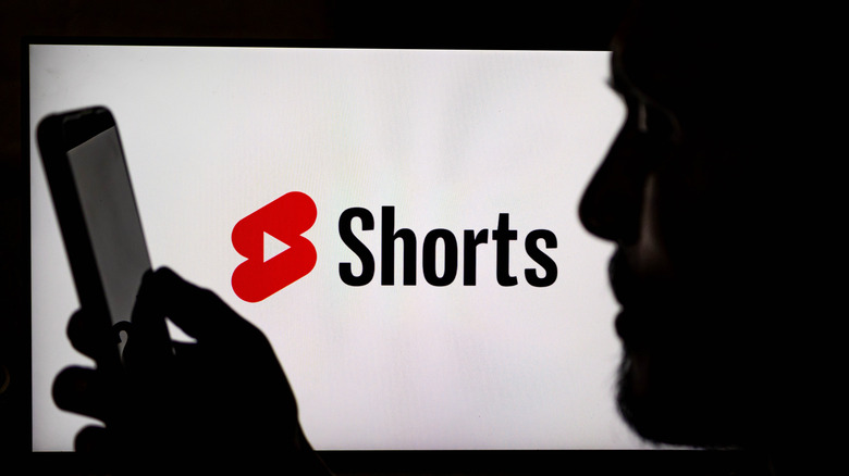 Imagen fotográfica de un hombre usando un teléfono inteligente con el logotipo de YouTube Shorts de fondo.