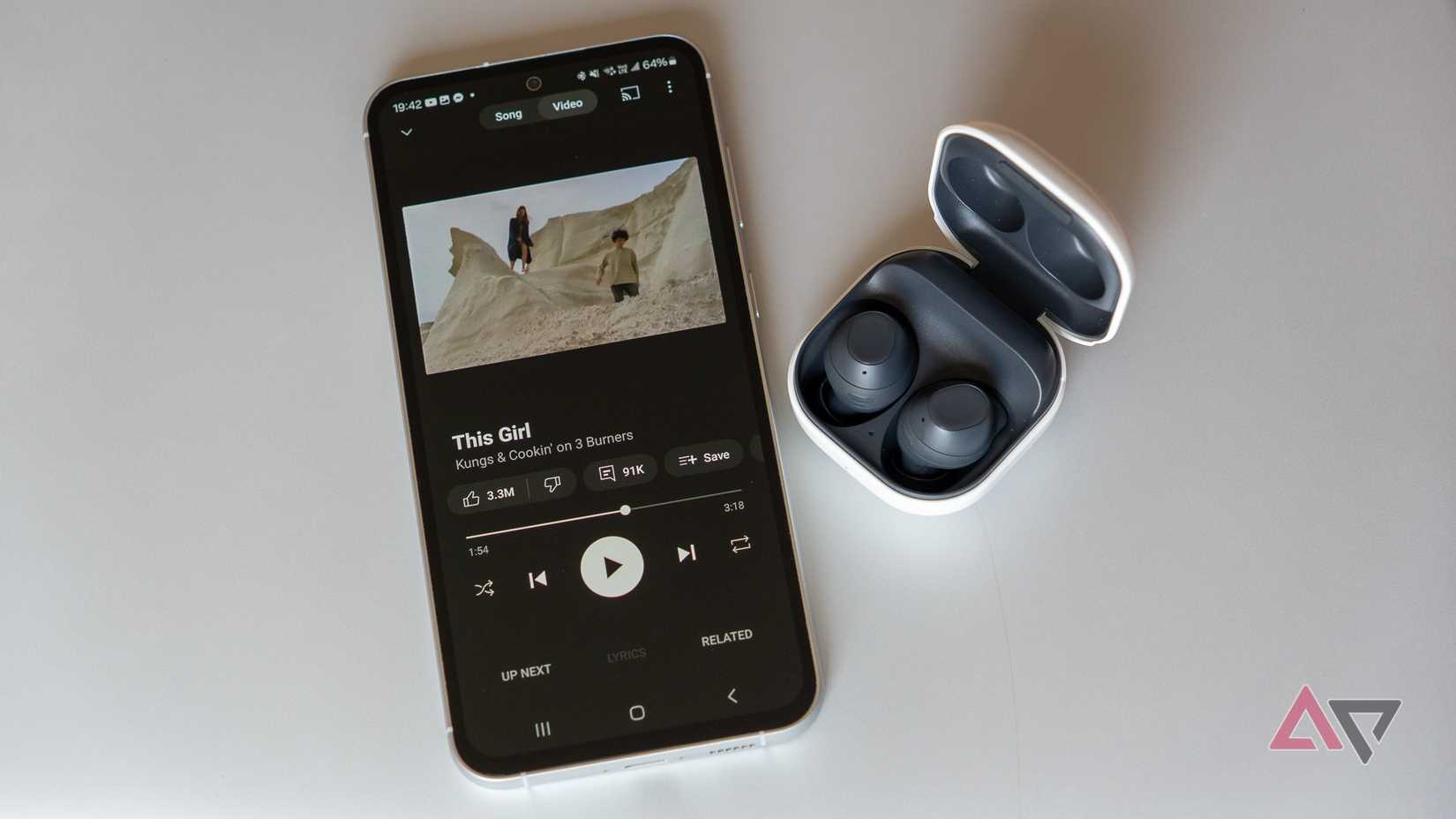 YouTube Music Premium se reproduce en un teléfono Samsung junto a unos auriculares inalámbricos Samsung sobre una superficie gris metalizada.